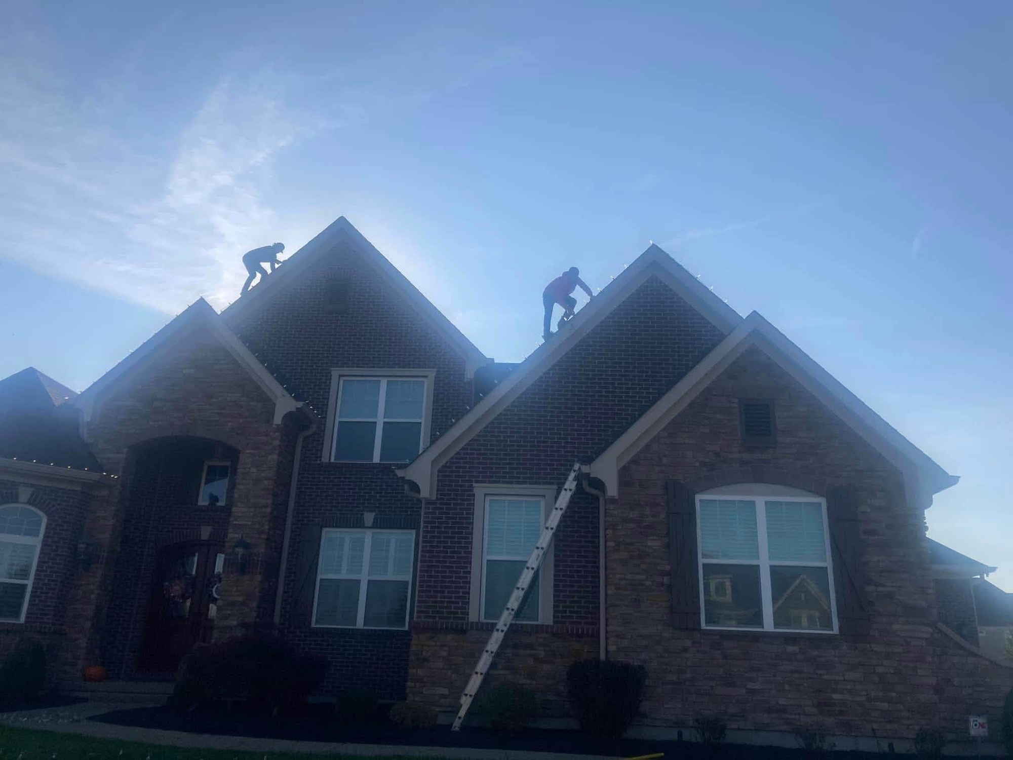 Two installers on ladders hanging Christmas lights along a roof ridge in Greater Cincinnati